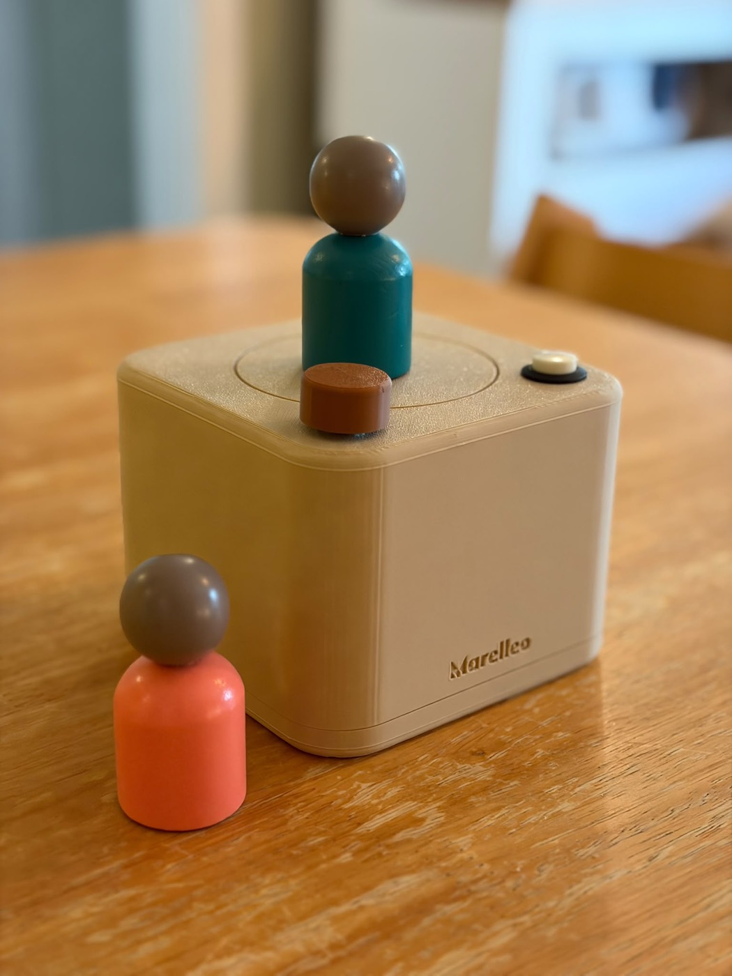 La boîte Marelleo terminée, un cube beige imprimé en 3D posé sur une table en bois, avec un cercle gravé sur le dessus. Deux figurines en bois peint, turquoise et corail, sont posées à côté et dessus.